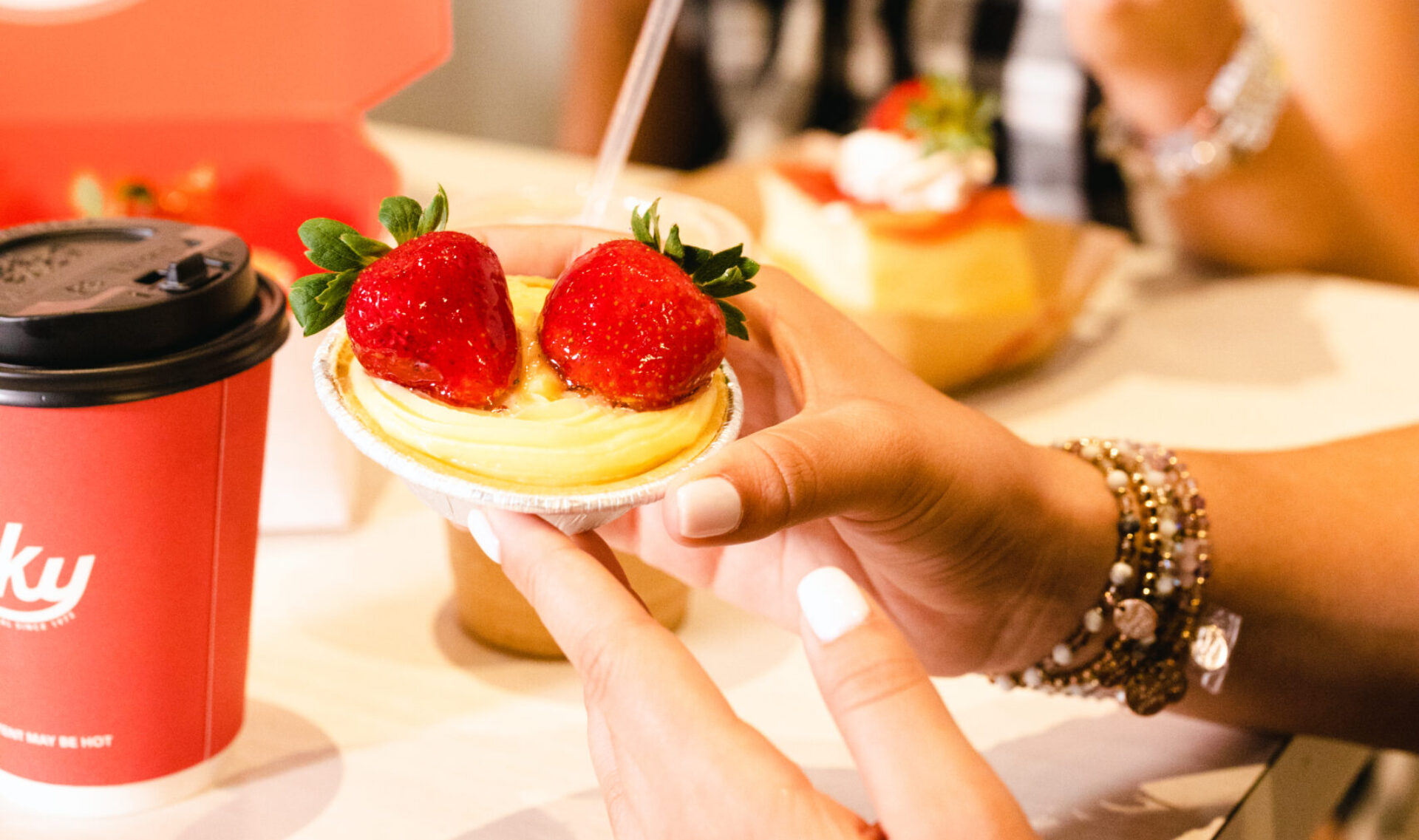 Close-up of a person holding a small fruit tart topped with glazed strawberries at Vicky Bakery, with a red branded coffee cup and other pastries visible on the table in the background