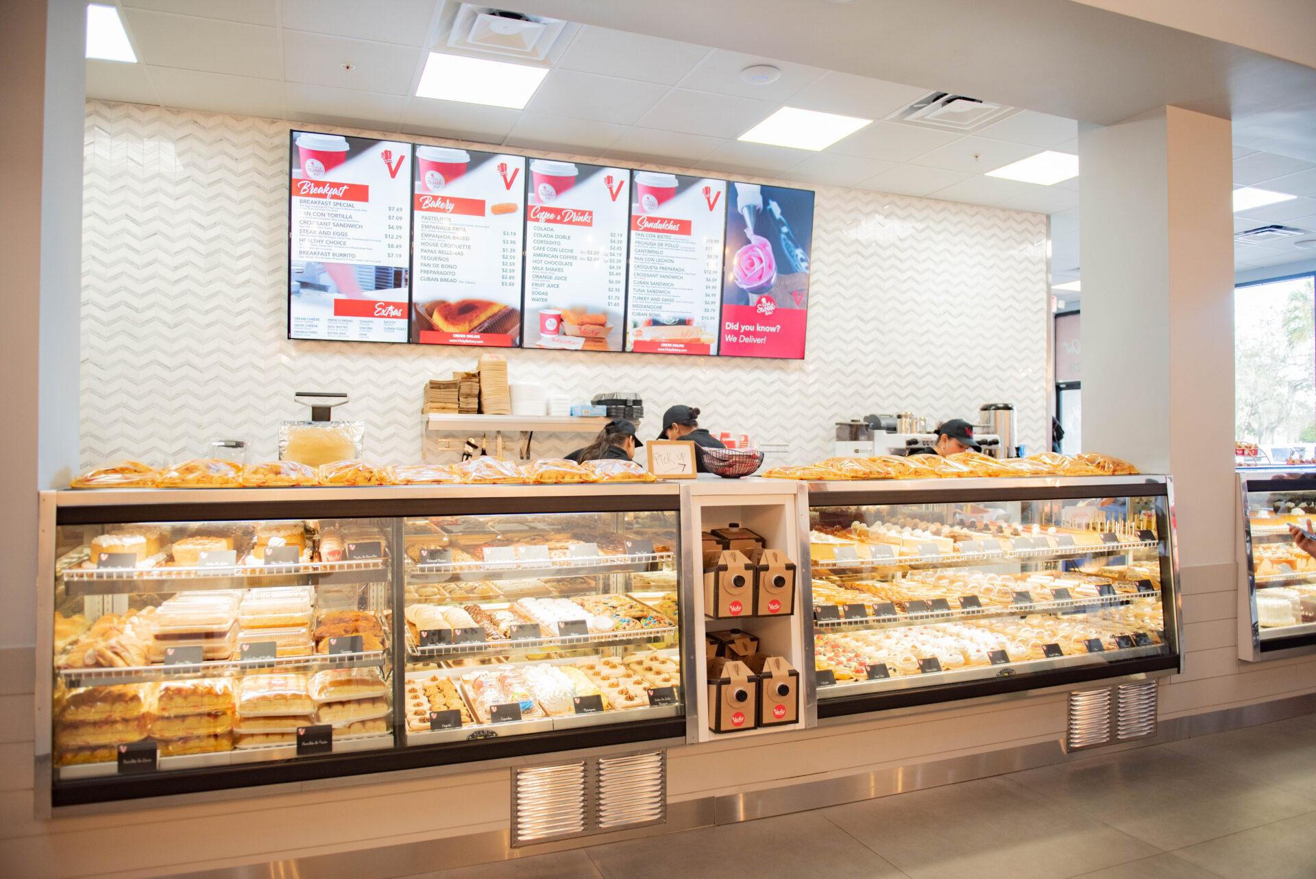 Interior of a Vicky Bakery location showing a well-lit pastry counter filled with baked goods, with overhead menu boards displaying items and prices on the back wall.