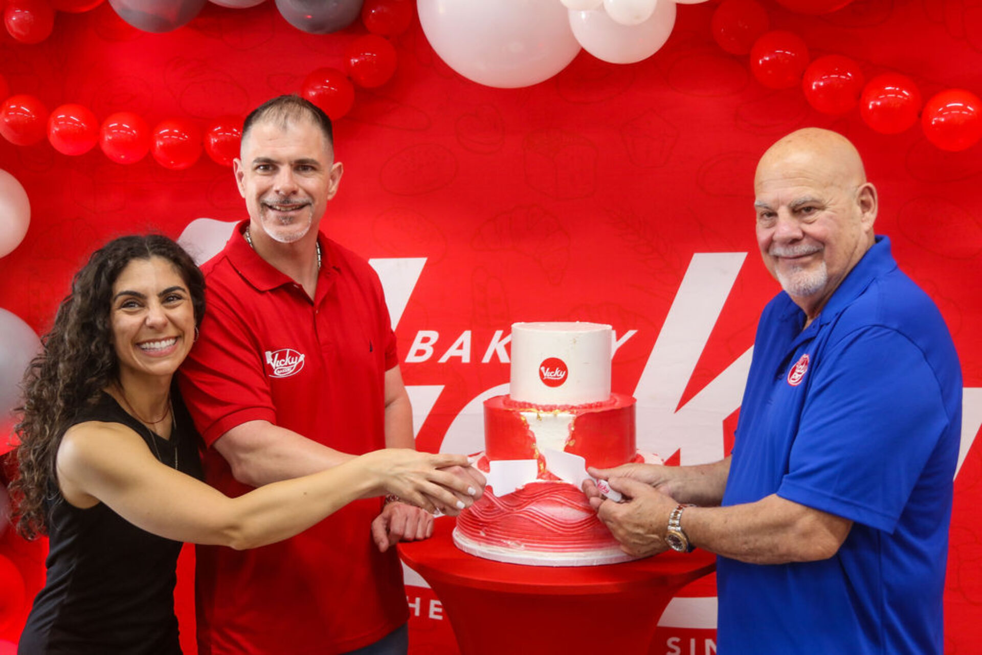 Three people smiling and cutting a red-and-white celebration cake at a Vicky Bakery event, standing in front of a branded red backdrop decorated with balloons.