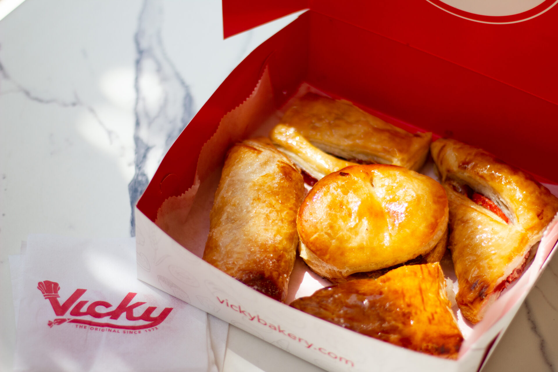 Box of assorted golden-brown Cuban pastries from Vicky Bakery, displayed on a marble surface with a branded napkin nearby.