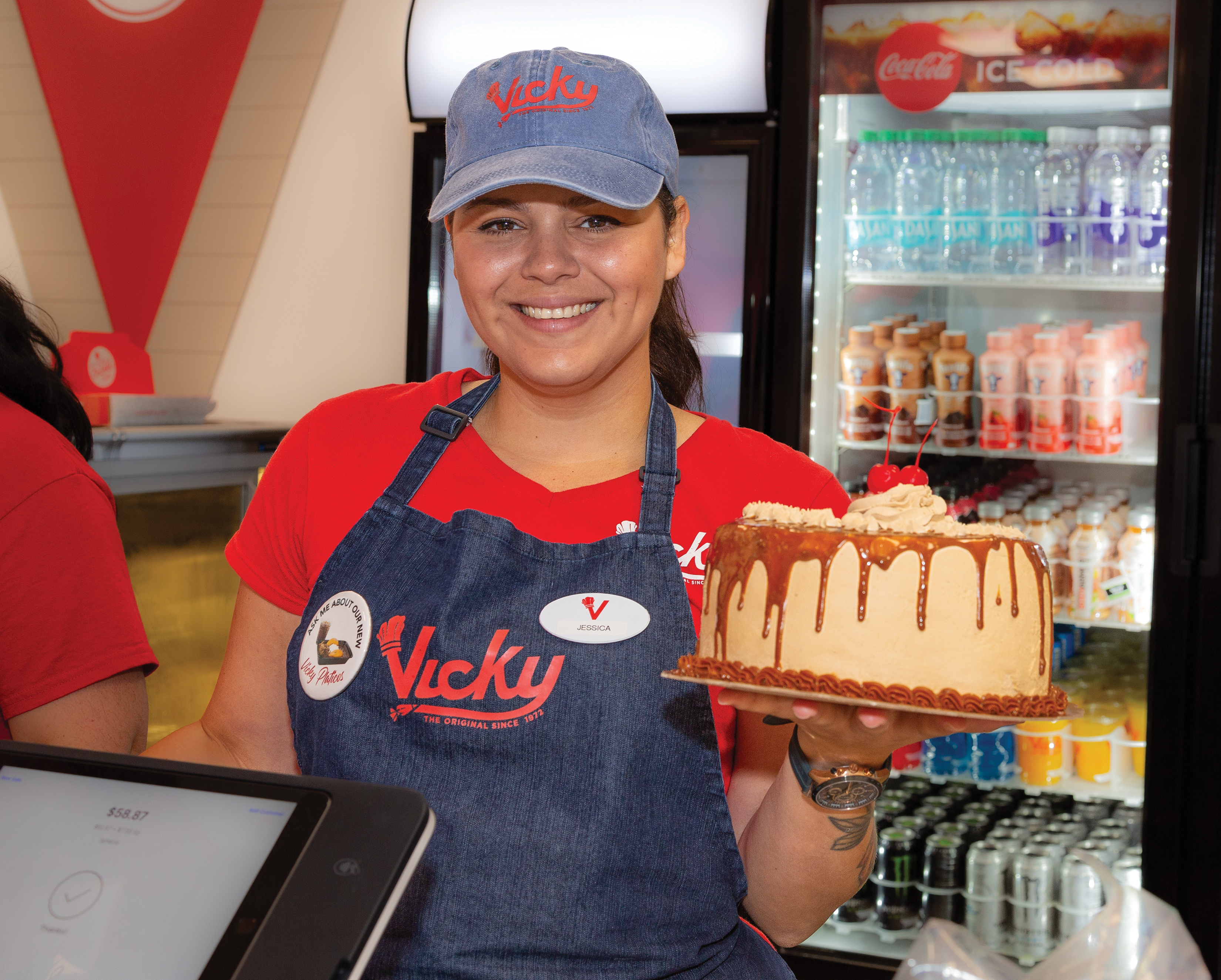 Smiling Vicky Bakery employee wearing a branded uniform and hat, holding a caramel-drizzled layer cake with whipped topping and a strawberry, inside a store near the register and beverage cooler.